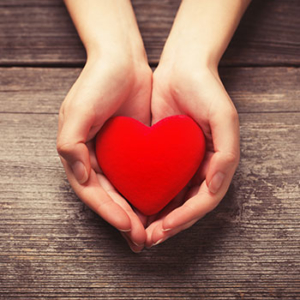 wooden background with hands coming from the top holding a small red heart in cupped hands