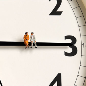 woman in orange dress and man in grey suit and hat sitting on the minutes hand of a large clock pointed at the 3 on the clock