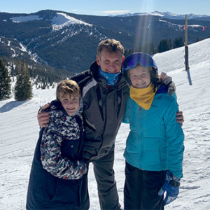 Financial Advisor Isaac Hartman with his two children in skiing outfits hugging and smiling in front of a view of the snow covered mountains